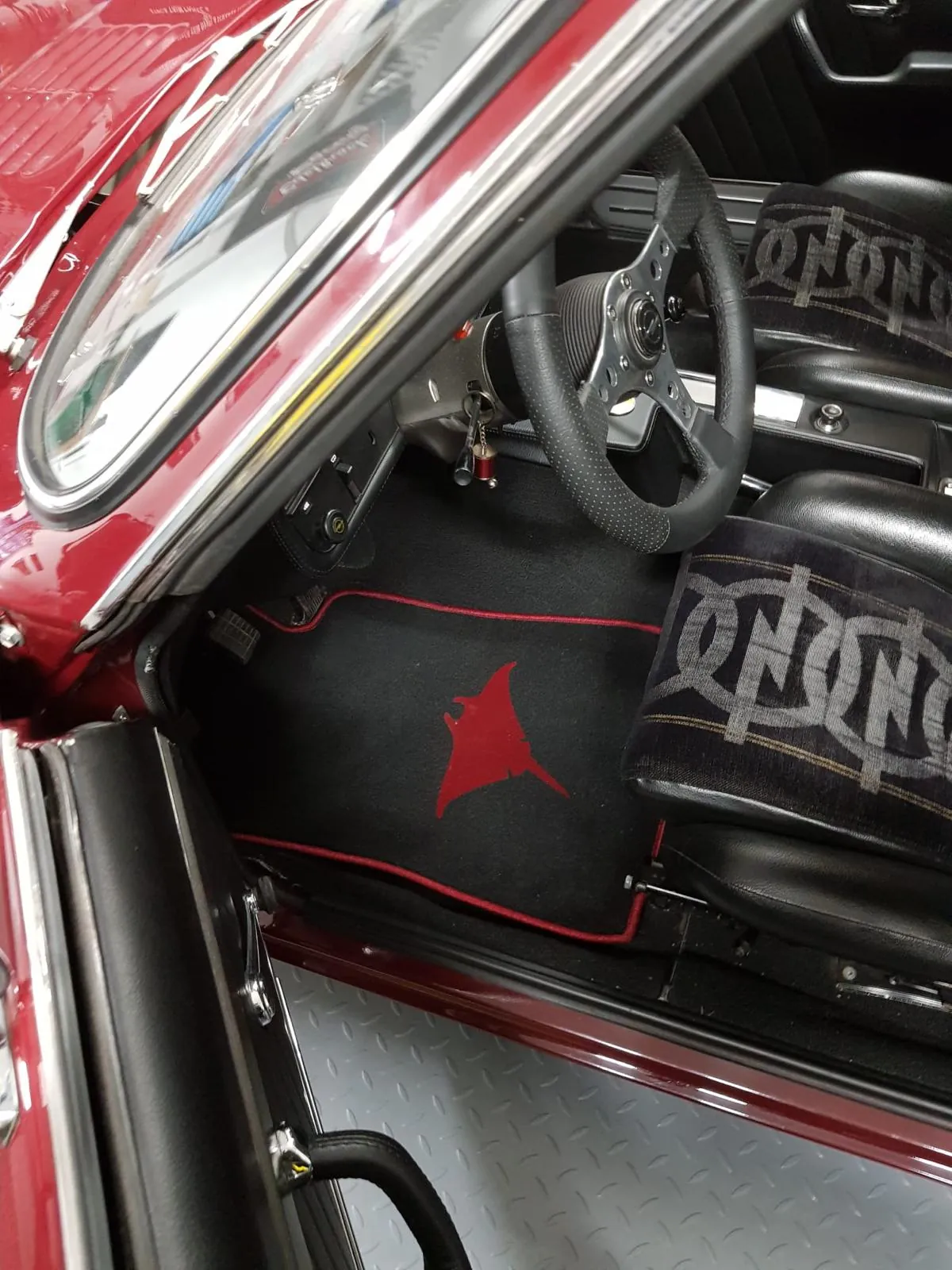 Blick in den Fahrerfußraum eines Opel Manta A, in dem eine schwarze Fußmatte mit rotem Rand liegt und einer roten Manta Motiv Bestickung