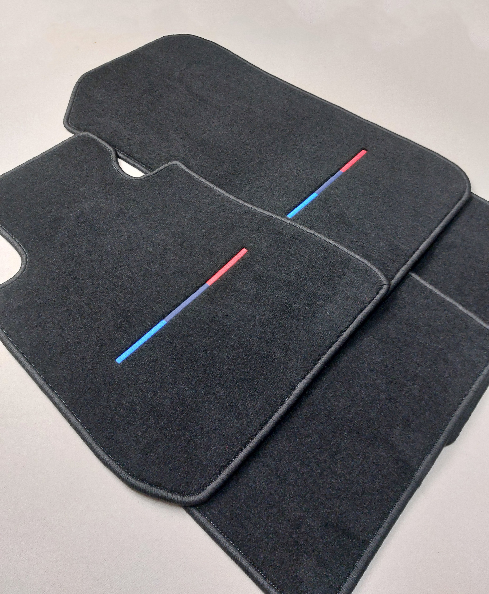4er Set M-Farbe seitlich Vier bestickte Fussmatten seitlich zu sehen,für einen BMW in den M Farben mittig. Der Teppich ist schwarz.